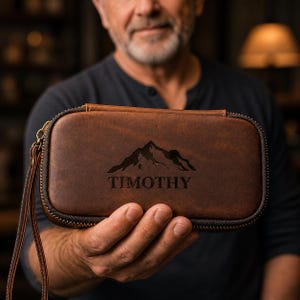 Puede incluir: Un estuche de cuero marr&oacute;n con un dise&ntilde;o de monta&ntilde;a y el nombre "TIMOTHY" grabado. El estuche tiene cierre de cremallera y una correa de mu&ntilde;eca. El estuche est&aacute; en la mano de una persona.
