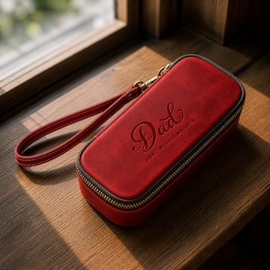 Puede incluir: Un estuche de cuero rojo con una correa de mu&ntilde;eca y cierre de cremallera. La palabra "Dad" est&aacute; grabada en relieve en la parte delantera, junto con los nombres "JAMES DAVID KAREN". El estuche es rectangular y parece un estuche o art&iacute;culo similar.