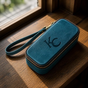 Puede incluir: Estuche con cremallera de color verde azulado con una correa de mu&ntilde;eca y herrajes dorados. El estuche presenta las iniciales "KC" en relieve en la parte delantera. La cremallera es marr&oacute;n y la correa es verde azulado con costuras a juego.