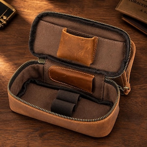 Puede incluir: Estuche de cuero marr&oacute;n abierto con cremallera, que revela un interior marr&oacute;n con bolsillos y bandas el&aacute;sticas. El estuche est&aacute; dise&ntilde;ado para guardar objetos peque&ntilde;os, con un aspecto robusto y texturizado.
