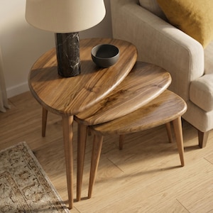 May include: Three wooden nesting tables with a natural finish and a distinctive, organic shape. The tables have tapered legs. A black marble lamp and a small grey bowl sit on the largest table. The tables are arranged in a living room setting.