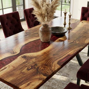 May include: A rectangular dining table featuring a natural wood slab surface with a central river inlay of deep red resin. The table is surrounded by four tufted burgundy velvet chairs and holds a ceramic vase with dried pampas grass.