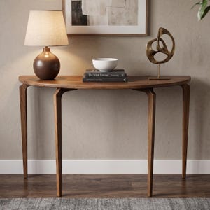 Op de afbeelding: Een halfronde houten sidetable met vier taps toelopende poten. Op de tafel staan een bruine lamp met een beige kap, een witte kom, twee op elkaar gestapelde boeken met de titel The Silent Listener en een bronzen abstract metalen beeld.