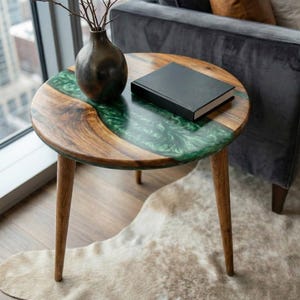 May include: Round wooden side table with a green epoxy resin inlay. The table has three wooden legs and a dark book on top. A dark vase with branches sits on the table. The table is approximately 24 inches in diameter.