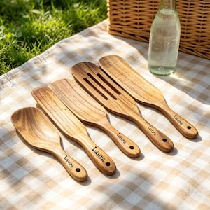 Peut inclure: Ensemble de cinq ustensiles de cuisine en bois, dont une cuillère, une spatule et une spatule ajourée, disposés sur une couverture de pique-nique à carreaux. Chaque ustensile porte le nom "Laura". Une bouteille d'eau en verre clair est également présente.