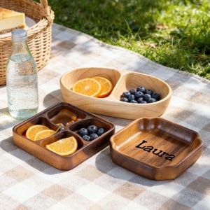 Può includere: Piatti di legno da portata pieni di frutta, tra cui fette d'arancia e mirtilli, sono disposti su una coperta da picnic. Un piatto è inciso con il nome "Laura". Una bottiglia d'acqua e un cestino sono sullo sfondo.