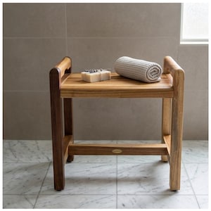 May include: A wooden shower bench with a light brown finish. A rolled gray waffle-weave towel and a bar of soap are on the bench. The bench has a rectangular seat and a lower shelf. The legs and frame are a darker brown.