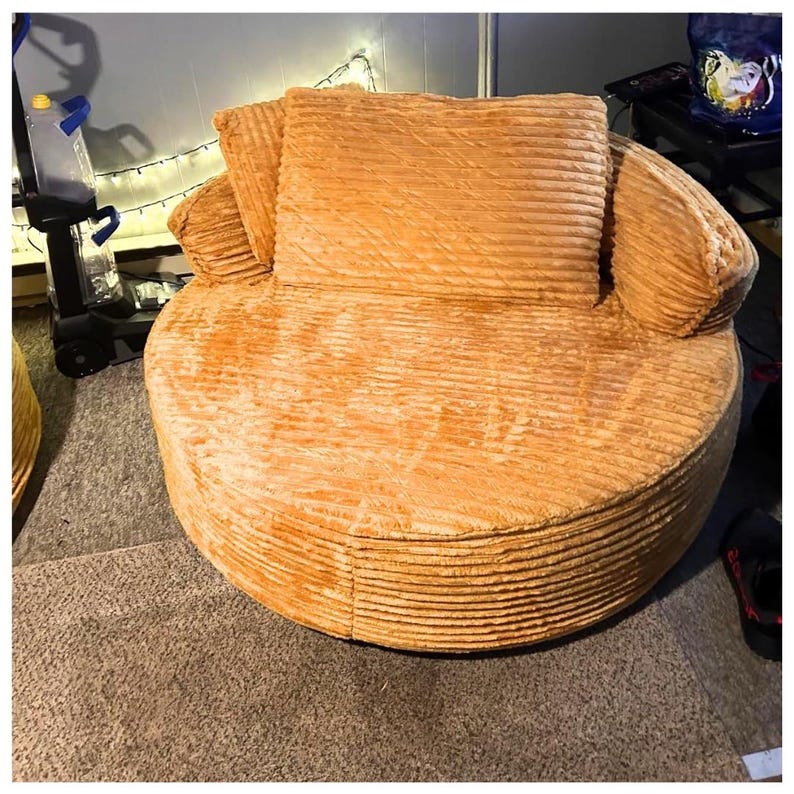 Puede incluir: Una silla grande y redonda con textura de pana en un tono naranja c&aacute;lido. La silla presenta un respaldo y reposabrazos redondeados, con un coj&iacute;n cuadrado grande y dos cojines m&aacute;s peque&ntilde;os. La silla est&aacute; sobre una alfombra de color neutro.