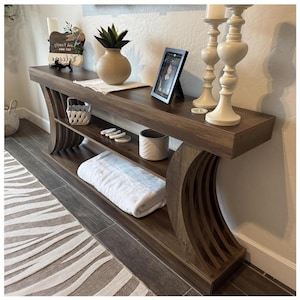 May include: A dark brown wooden console table with curved side supports. The table has two shelves, displaying a white towel, a small basket, and decorative items. A framed photo, candles, and a vase with a plant are on top. The rug has a zebra stripe pattern.