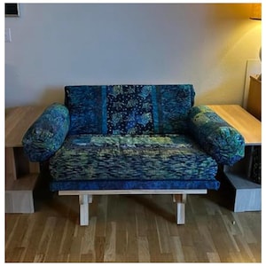May include: A futon sofa with a wooden frame and a patterned fabric in shades of blue and green. The sofa has two cylindrical armrests and a backrest, all covered in the same fabric. The cushions have a quilted appearance.
