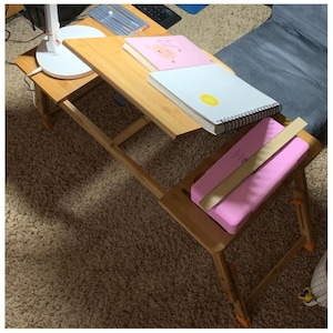 May include: A bamboo laptop desk with a tilting surface and folding legs. The desk holds a white spiral-bound notebook with a yellow circle, a pink notebook with a pig illustration, and a pink cushion with a wooden ruler.