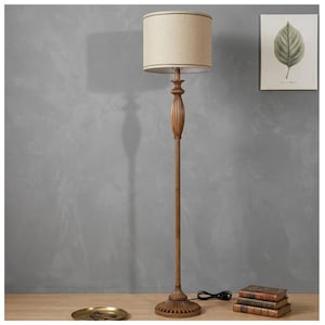May include: A tall, antique-style floor lamp with a beige fabric shade and a brown, ornate wooden base. The lamp is positioned on a wooden surface, with a small brass tray and a stack of books nearby. The background is a gray wall.