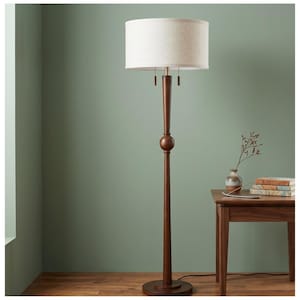 May include: A tall, wooden floor lamp with a beige drum shade. The lamp has a dark brown finish and a round base. The lamp is next to a small wooden table with a vase and books.
