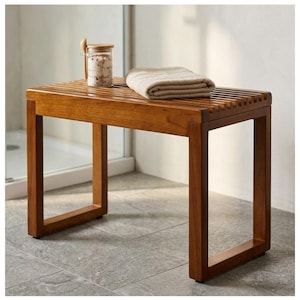 May include: A wooden shower bench with a slatted top and square legs. A folded beige towel and a glass jar with a wooden spoon rest on the bench. The bench is made of a warm-toned wood.