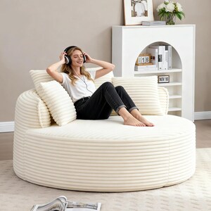 Puede incluir: Un sof&aacute; redondo de pana color crema con cojines a juego. Una persona con auriculares est&aacute; sentada en el sof&aacute;. Una estanter&iacute;a blanca con libros y objetos decorativos se encuentra en el fondo.