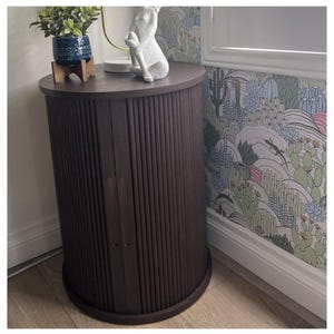 May include: A dark brown, cylindrical cabinet with vertical wooden slats. The cabinet has a rounded top and base. A small blue and white potted plant sits on top, along with a white statue. The cabinet is placed next to a wall with a desert-themed wallpaper.