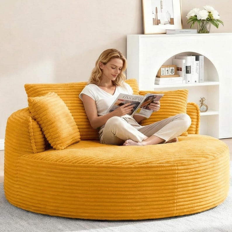 Puede incluir: Un sof&aacute; redondo de pana amarillo mostaza con dos cojines a juego y un respaldo. El sof&aacute; es una opci&oacute;n de asiento c&oacute;moda para una sala de estar o zona de descanso. La imagen muestra a una persona sentada en el sof&aacute; leyendo una revista.