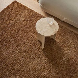May include: A brown textured rug with a small round side table and a glass of water. The rug is a solid color and appears to be made of a woven material. The table is made of a light-colored stone.