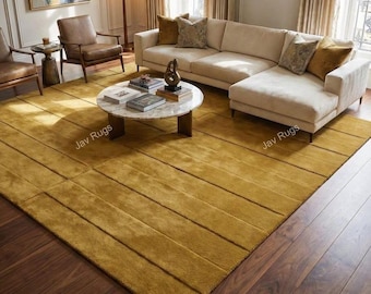 May include: A mustard yellow rectangular rug with a textured, linear design. The rug is placed on a dark wood floor, with a cream-colored sectional sofa and a marble coffee table in the background. The rug is a home decor item.