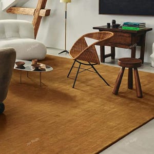 May include: A room with a large, rectangular, golden-brown rug. The room includes a white curved armchair, a woven chair, a small wooden stool, and a dark wooden table. A small, oval coffee table with a glass top and metal legs is also present.