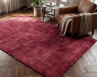 May include: A large, rectangular rug in a deep burgundy colour. The rug has a soft, plush texture and is placed on a dark wood floor. The rug is in a living room setting, with a leather armchair and side table visible.