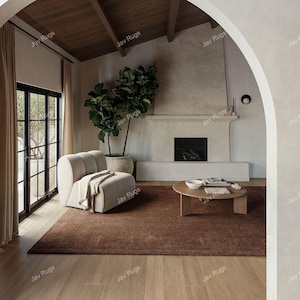 Puede incluir: Un salón moderno con una alfombra marrón, un sillón beige y una mesa de centro de madera. Una gran planta en maceta y una chimenea contribuyen a la atmósfera acogedora. La habitación tiene suelos de madera y un techo alto.