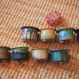May include: A collection of small, handcrafted ceramic pots in various colors, including blue, green, and tan. Each pot has a unique glaze and sits on a textured surface. A succulent plant is also present.