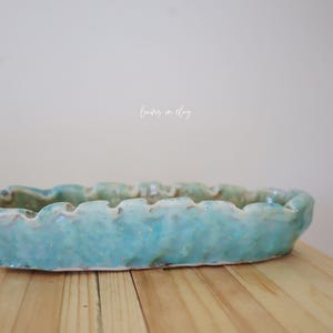 May include: A rectangular, turquoise ceramic dish with a wavy rim. The handmade pottery has a textured surface and a light blue glaze. The dish is displayed on a wooden surface.