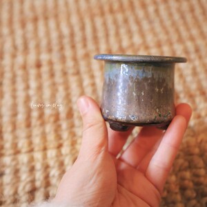 May include: A small, cylindrical ceramic pot with a wide, flat rim and three short, stubby feet. The glaze features a gradient of muted blue at the top transitioning into a textured, earthy brown and grey finish. The text "leaves in clay" is visible.