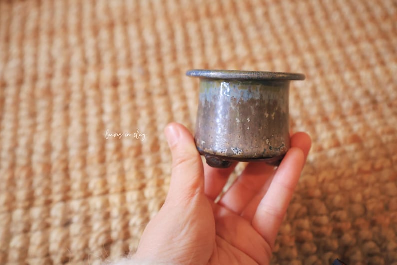 May include: A small, cylindrical ceramic pot with a wide, flat rim and three short, stubby feet. The glaze features a gradient of muted blue at the top transitioning into a textured, earthy brown and grey finish. The text "leaves in clay" is visible.