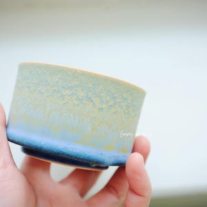 May include: A handmade ceramic cup with a cylindrical shape. The cup features a gradient of colors, transitioning from a deep blue base to a light blue and yellow rim. The cup is held in a hand.