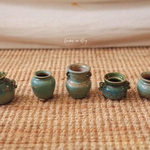 May include: Five small, handcrafted ceramic vases in varying shades of teal and green are arranged in a row. The vases have a rustic, textured appearance, with some featuring small handles. The background is a neutral beige.