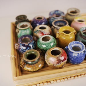 May include: A collection of small, round ceramic pots with various colorful glazes, including blue, green, yellow, and brown, arranged on a bamboo tray. The text "Leaves in Clay" is visible on the left side of the tray, which features a slatted design.