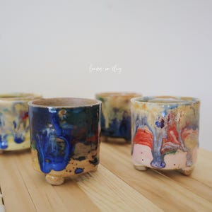May include: Four handmade ceramic cups with a unique, colorful glaze. The cups are cylindrical with small, rounded feet. The glaze features a mix of blue, yellow, red, and brown hues, creating a dripped effect. The cups are arranged on a light wooden surface.