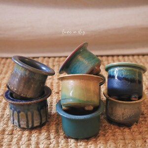 May include: A collection of small ceramic pots with varied glaze finishes in shades of blue, green, and tan. The pots feature wide rims and short feet, with some displaying drip patterns. The text "leaves in clay" is visible in the center.