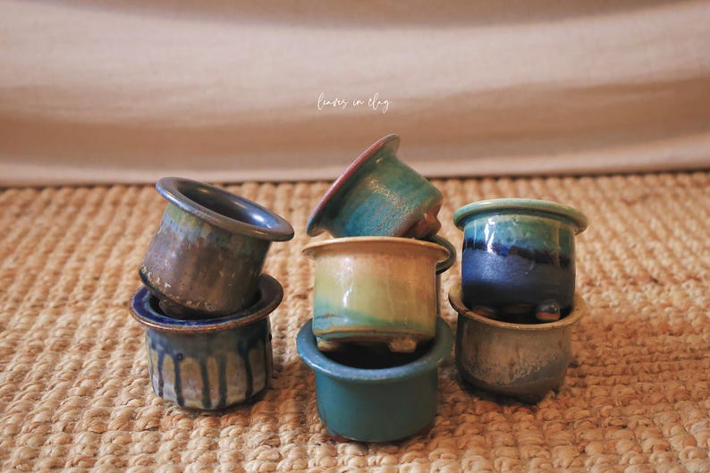 May include: A collection of small ceramic pots with varied glaze finishes in shades of blue, green, and tan. The pots feature wide rims and short feet, with some displaying drip patterns. The text "leaves in clay" is visible in the center.