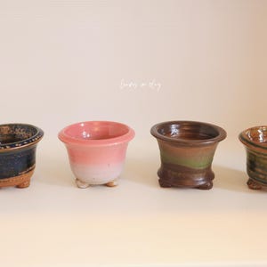 May include: Four small ceramic pots with three feet each, arranged in a row. The pots feature distinct glazes: one dark blue with brown, one pink fading to white, one brown with a green band, and one brown with green horizontal stripes.