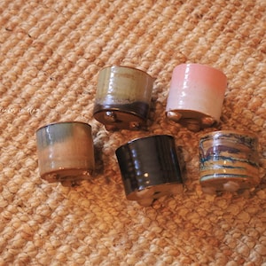 May include: Five small ceramic pots in various colors, including pink, brown, and blue, each with a unique glaze. The pots have a cylindrical shape and are elevated on small feet. The pots are arranged on a textured, woven surface.