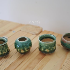 May include: Four small, handcrafted ceramic vessels with a green and yellow glaze. The vessels are arranged on a light-colored wooden surface. One has a handle, and another has a flared rim. The text "lovers in clay" is visible.