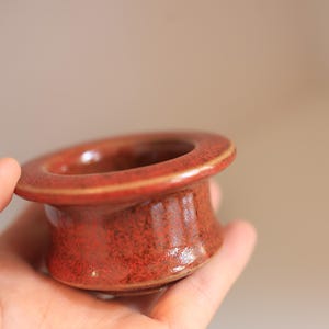 May include: A small, round ceramic vessel with a wide, flat rim and a tapered base. The object features a mottled, deep red glaze with subtle brown speckles throughout, creating a textured appearance across the entire exterior surface.