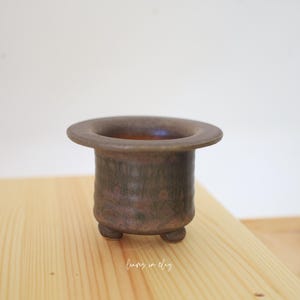 May include: A small, cylindrical ceramic planter with a wide, flat rim and three short, rounded feet. The exterior features a mottled, dark brown and earthy bronze glaze with a textured, matte finish. The interior is smooth and dark.