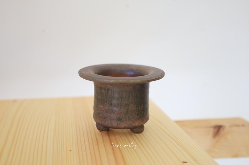 May include: A small, cylindrical ceramic planter with a wide, flat rim and three short, rounded feet. The exterior features a mottled, dark brown and earthy bronze glaze with a textured, matte finish. The interior is smooth and dark.