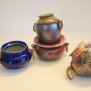 May include: A collection of small ceramic pots in various colors. One is deep blue, another is a mix of brown and red, and the third is a combination of brown, red, and blue. Each pot has brass ring handles.