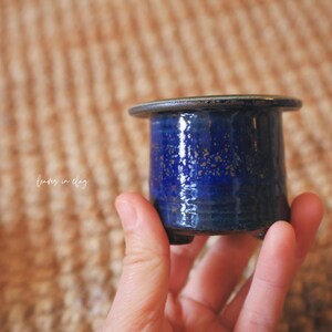 May include: A small, cylindrical ceramic pot with a glossy, deep blue glaze featuring subtle gold-toned speckles. The vessel has a wide, flat rim, a smooth body, and small feet at the base. The text "leaves in clay" is visible on the side.