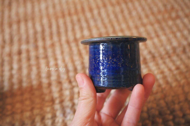 May include: A small, cylindrical ceramic pot with a glossy, deep blue glaze featuring subtle gold-toned speckles. The vessel has a wide, flat rim, a smooth body, and small feet at the base. The text "leaves in clay" is visible on the side.