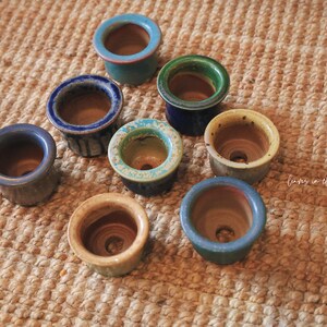 May include: A collection of eight small, round ceramic pots with drainage holes, featuring various glazed finishes in shades of blue, green, and yellow. Each pot has a smooth, wide rim and a natural clay interior, arranged in a cluster.