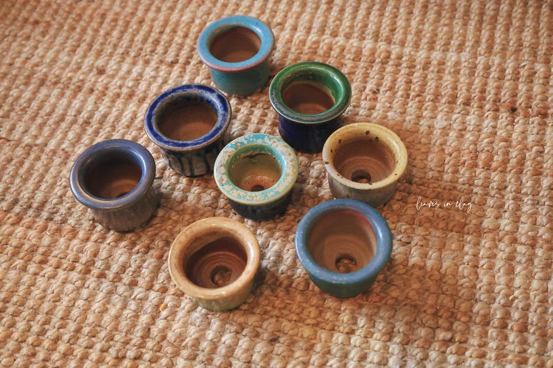 May include: A collection of eight small, round ceramic pots with drainage holes, featuring various glazed finishes in shades of blue, green, and yellow. Each pot has a smooth, wide rim and a natural clay interior, arranged in a cluster.