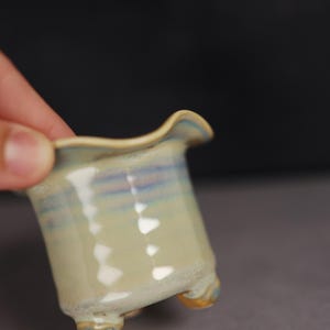 May include: A small ceramic planter with a wavy, flared rim and three short feet. The exterior features a glossy, light cream-colored glaze with horizontal bands of soft blue and purple hues, creating a subtle, multi-toned appearance.