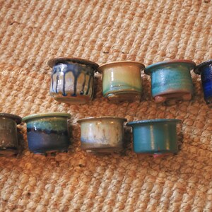 May include: Eight small ceramic pots arranged in two rows, each featuring a distinct glazed finish in shades of blue, green, brown, and tan. These cylindrical containers have wide rims and small feet, showcasing varied drip and solid glaze patterns.