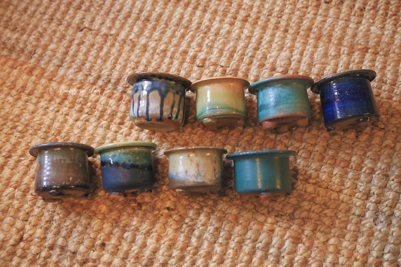 May include: Eight small ceramic pots arranged in two rows, each featuring a distinct glazed finish in shades of blue, green, brown, and tan. These cylindrical containers have wide rims and small feet, showcasing varied drip and solid glaze patterns.